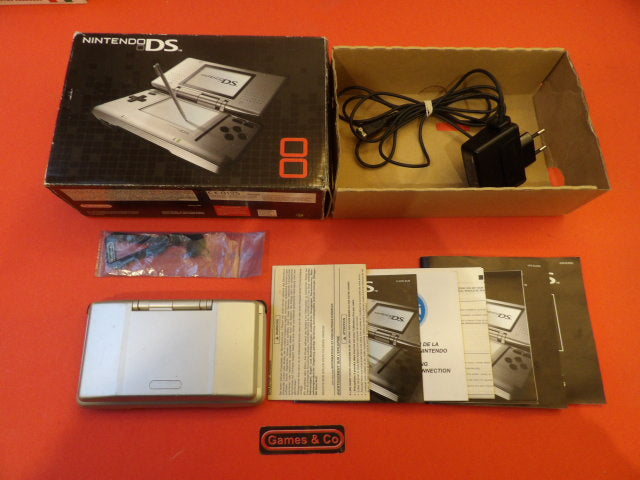Nintendo DS console with charger in box, including manuals and stylus, on a bright orange background.
